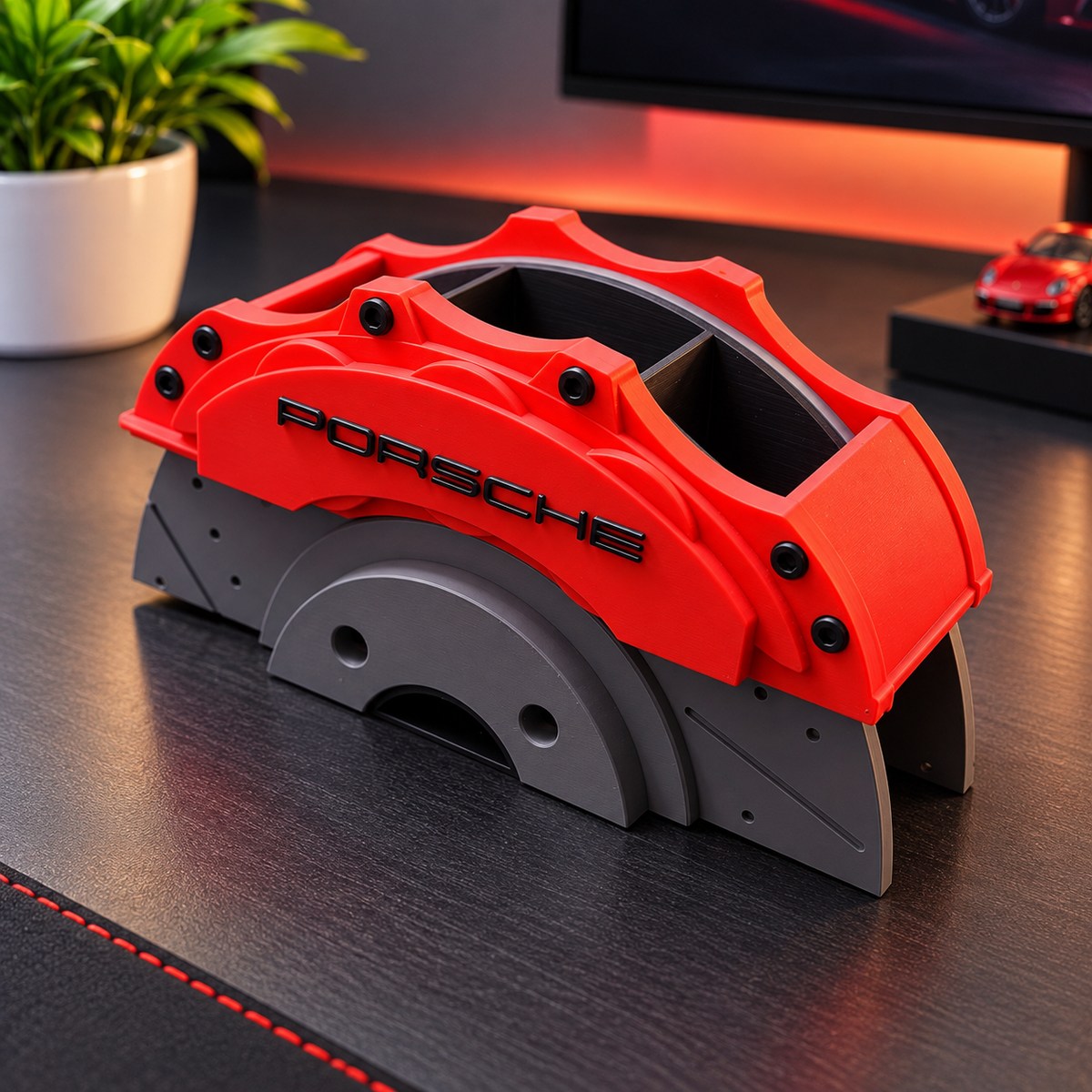 A red and gray Porsche-branded decorative object or model part displayed on a dark desk, with a plant, monitor, and small car model in the background.