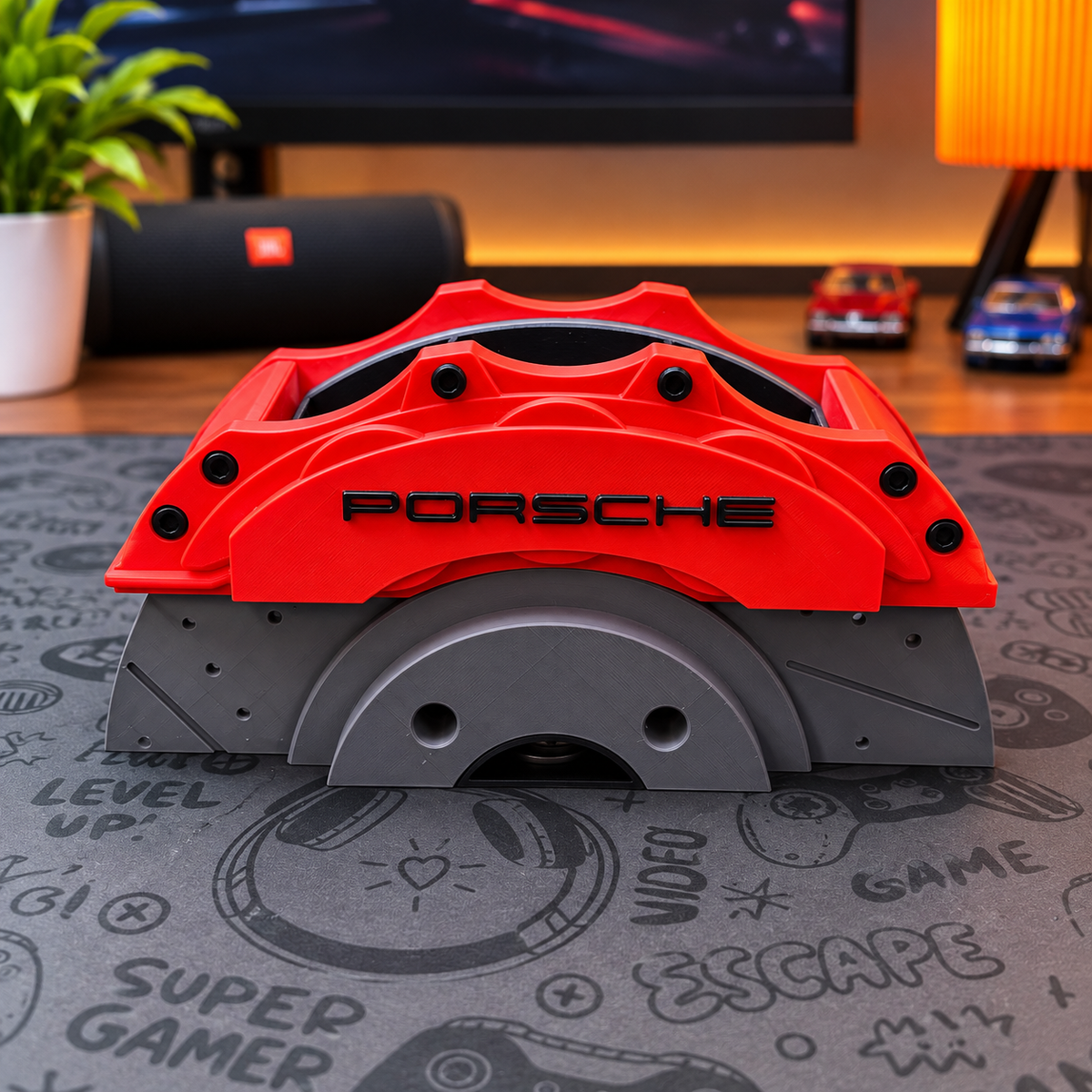 A red Porsche-branded brake caliper replica or decorative prop with black lettering, displayed on a gray gaming mat in a room setting with a speaker, plant, and desk items in the background.