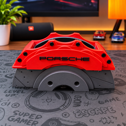 A red Porsche-branded brake caliper replica or decorative prop with black lettering, displayed on a gray gaming mat in a room setting with a speaker, plant, and desk items in the background.