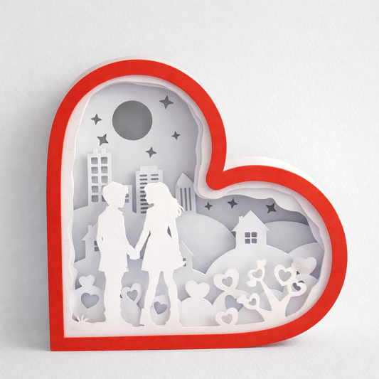 Heart-shaped paper cutout with a couple and cityscape inside, on a light gray background